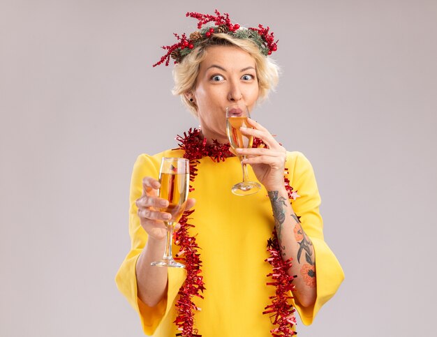 Impressed young blonde woman wearing christmas head wreath and tinsel garland around neck holding two glasses of champagne looking at camera drinking one isolated on white background