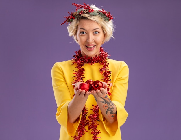 Free Photo impressed young blonde woman wearing christmas head wreath and tinsel garland around neck looking  holding christmas baubles isolated on purple wall with copy space