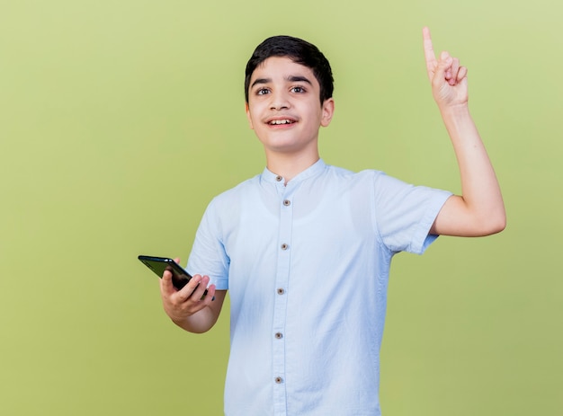 Impressed young boy holding mobile phone looking at front pointing up isolated on olive green wall