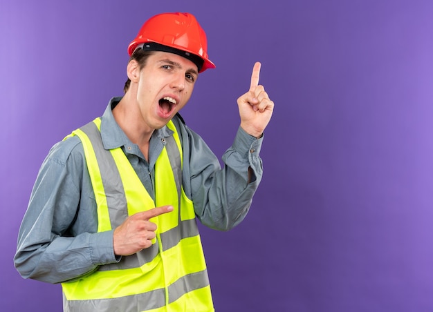 Impressed young builder man in uniform points at side isolated on blue wall with copy space