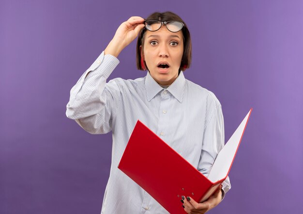 Impressed young call center girl wearing glasses holding folder and glasses isolated on purple 