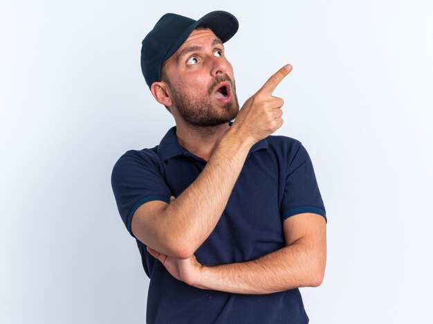 Impressed young caucasian delivery man in blue uniform and cap looking and pointing up isolated on white wall