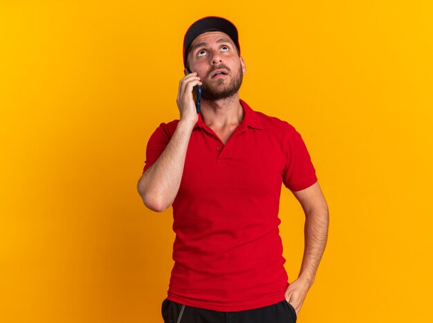 Impressed young caucasian delivery man in red uniform and cap keeping hand in pocket talking on phone looking up isolated on orange wall with copy space