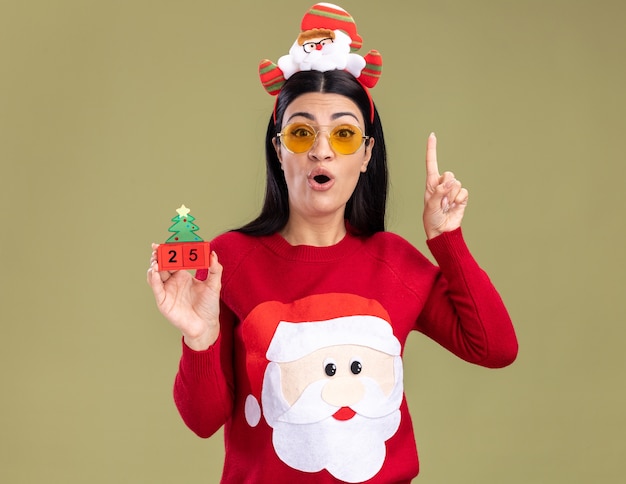 Free photo impressed young caucasian girl wearing santa claus headband and sweater with glasses holding christmas tree toy with date  pointing up isolated on olive green wall