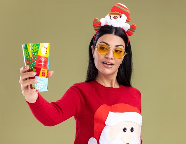 Impressed young caucasian girl wearing santa claus headband and sweater with glasses stretching out plastic christmas cup looking at it isolated on olive green background