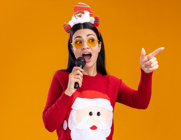 Impressed young caucasian girl wearing santa claus headband and sweater with glasses talking into microphone looking and pointing at side isolated on orange background