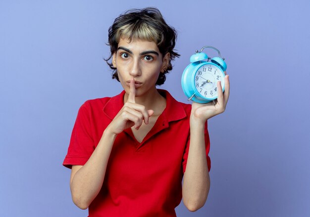 Impressed young caucasian girl with pixie haircut holding alarm clock gesturing silence isolated on purple background with copy space