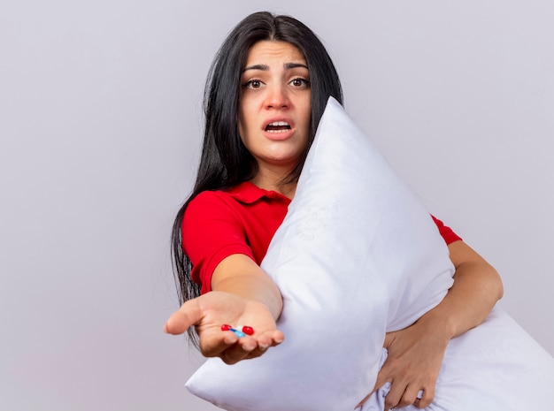 Free photo impressed young caucasian ill girl hugging pillow looking at camera stretching out capsules towards camera isolated on white background with copy space