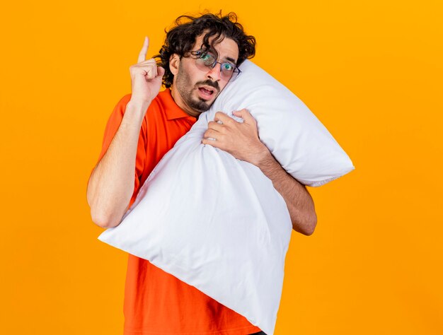 Impressed young caucasian ill man wearing glasses hugging pillow looking at camera raising finger isolated on orange background with copy space