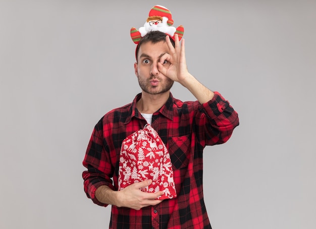 Impressed young caucasian man wearing christmas headband holding christmas sack looking at camera doing look gesture with pursed lips isolated on white background