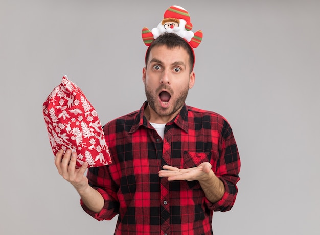 Impressed young caucasian man wearing christmas headband holding and pointing with hand at christmas sack  isolated on white wall with copy space