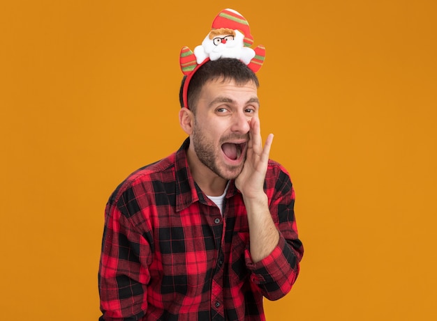 Impressed young caucasian man wearing santa claus headband  keeping hand near mouth whispering isolated on orange wall with copy space