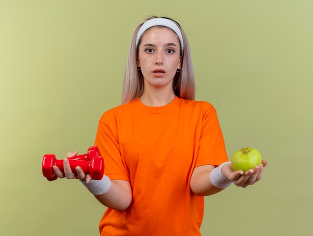 Free photo impressed young caucasian sporty girl with braces wearing headband and wristbands holds dumbbells and apple