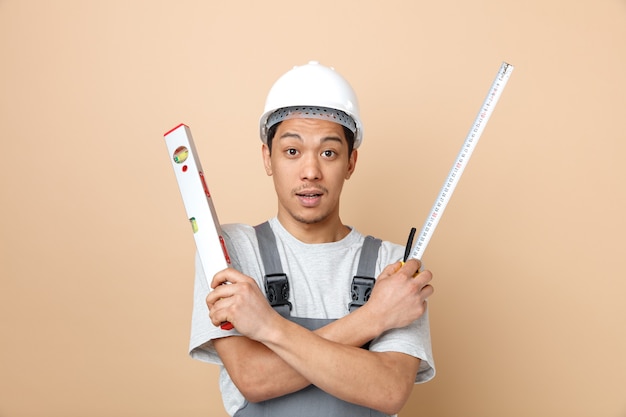 Impressed young construction worker wearing safety helmet and uniform holding tape meter and level ruler keeping hands crossed 