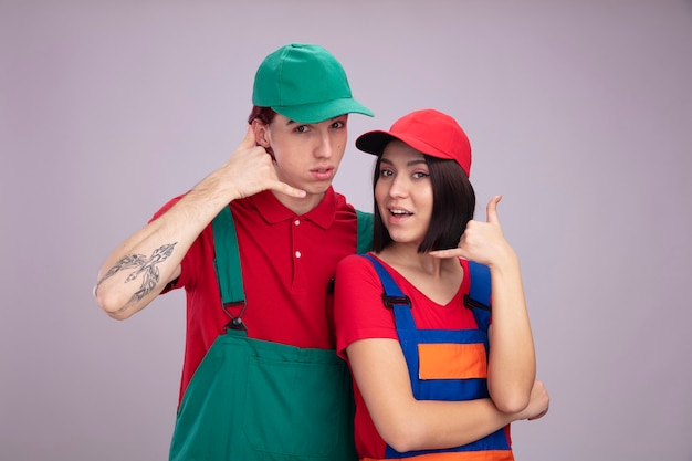 Free Photo impressed young couple in construction worker uniform and cap looking at camera doing call gesture isolated on white wall