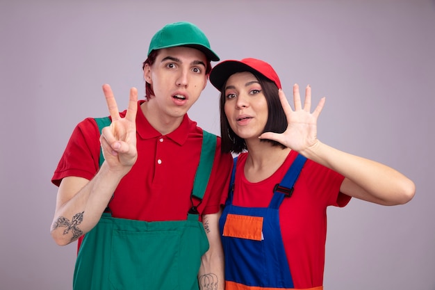 Impressed young couple in construction worker uniform and cap looking at camera guy showing two with hand girl showing five with hand isolated on white wall