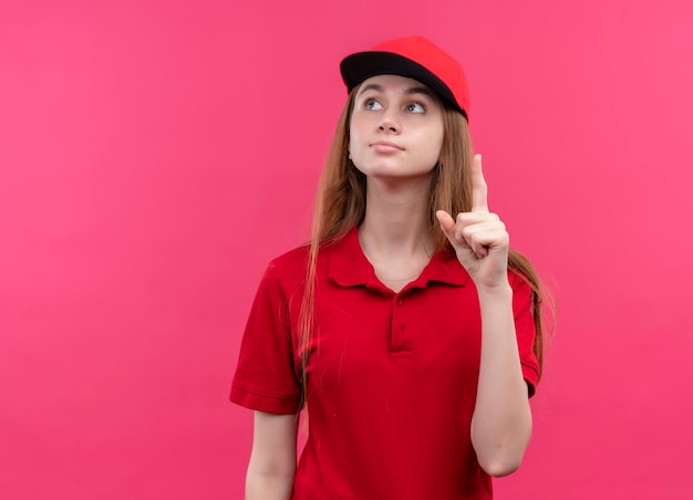 Impressed young delivery girl in red uniform looking up with raised finger on isolated pink space with copy space