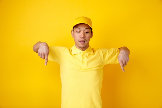 Impressed young delivery man wearing cap and uniform looking and pointing down 