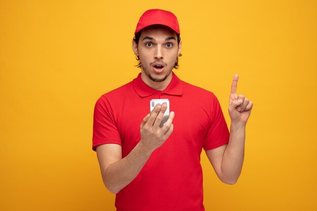 impressed young delivery man wearing uniform and cap holding mobile phone looking at camera pointing up isolated on yellow background