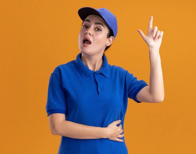 Impressed young delivery woman in uniform and cap pointing up