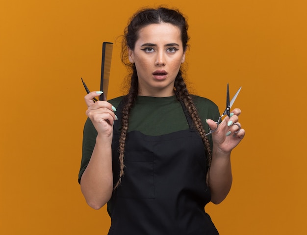 Impressed young female barber in uniform holding barber tools isolated on orange wall with copy space