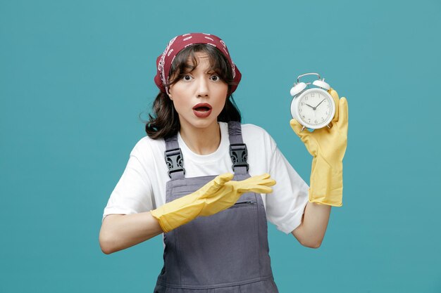 Impressed young female cleaner wearing uniform bandana and rubber gloves showing alarm clock looking at camera pointing with hand at clock isolated on blue background