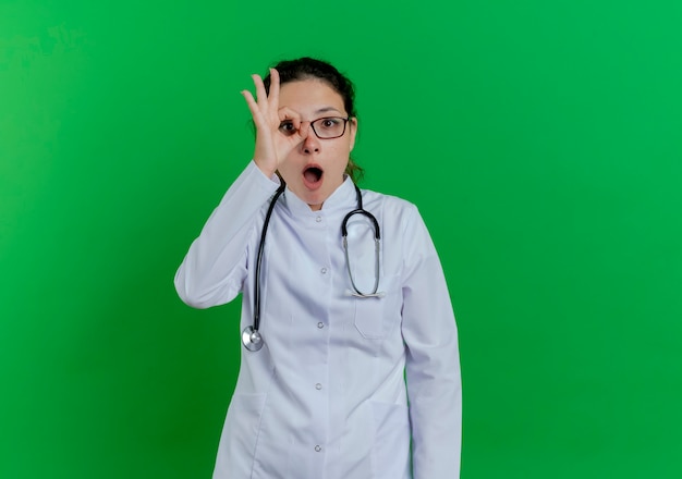 Impressed young female doctor wearing medical robe and stethoscope and glasses  doing look gesture isolated on green wall with copy space