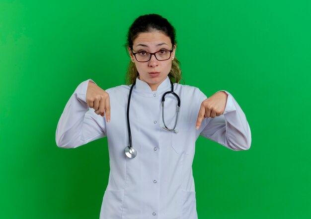 Impressed young female doctor wearing medical robe and stethoscope and glasses  pointing down isolated on green wall