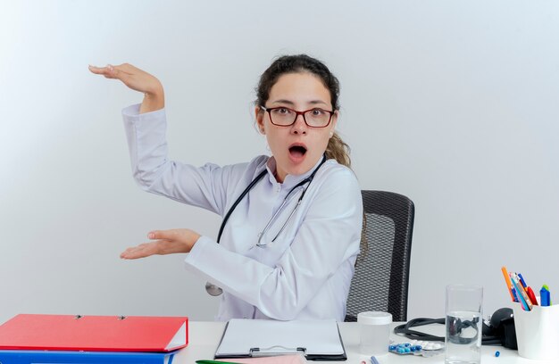 Impressed young female doctor wearing medical robe and stethoscope and glasses sitting at desk with medical tools looking doing size gesture isolated