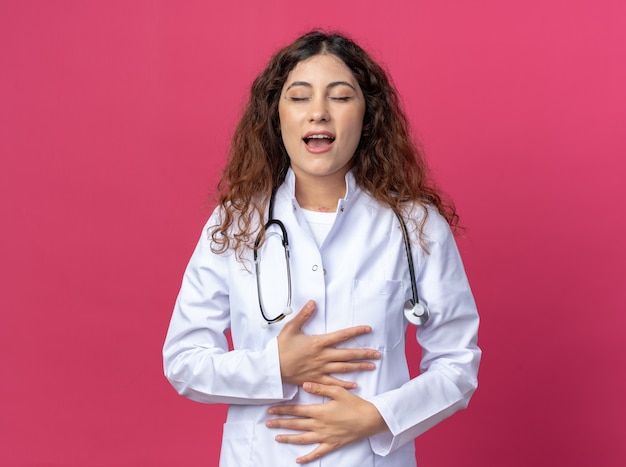 Impressed young female doctor wearing medical robe and stethoscope keeping hands on belly with closed eyes isolated on pink wall