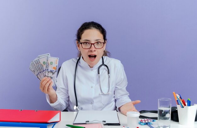 Impressed young female doctor wearing medical robe and stethoscope sitting at desk with medical tools holding money looking isolated