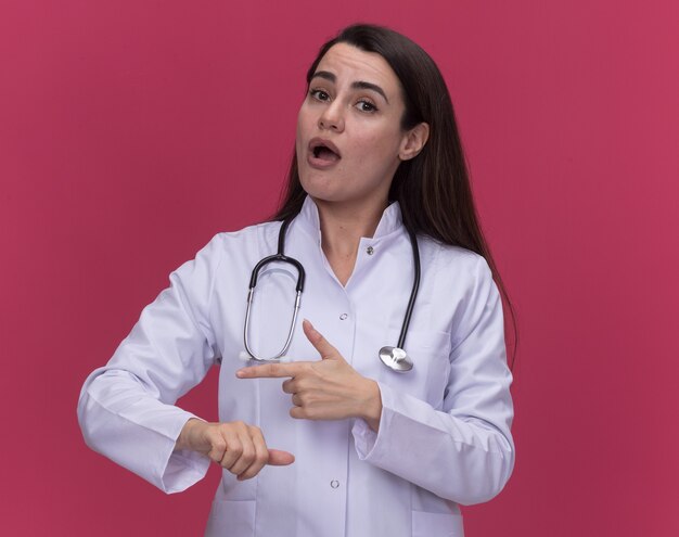 Impressed young female doctor wearing medical robe with stethoscope points at sides on pink 