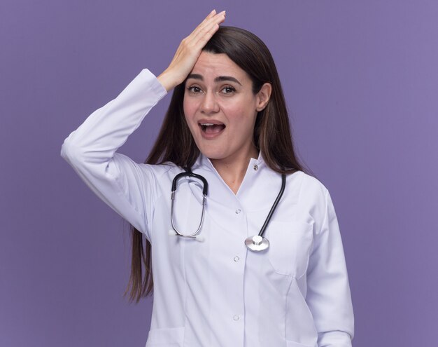 Impressed young female doctor wearing medical robe with stethoscope puts hand on head and looks at camera on purple 