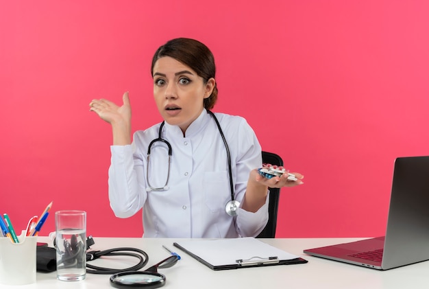 Impressed young female doctor wearing medical robe with stethoscope sitting at desk work on computer with medical tools holding pills and points with hand to side on pink wall with copy space