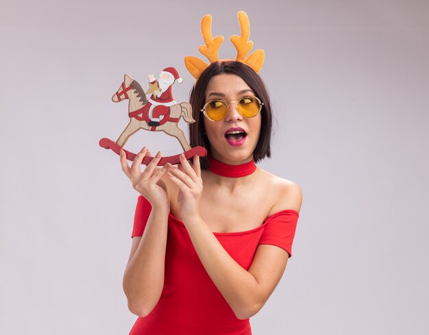 Impressed young girl wearing reindeer antlers headband and glasses holding and looking at santa on rocking horse figurine isolated on white background with copy space