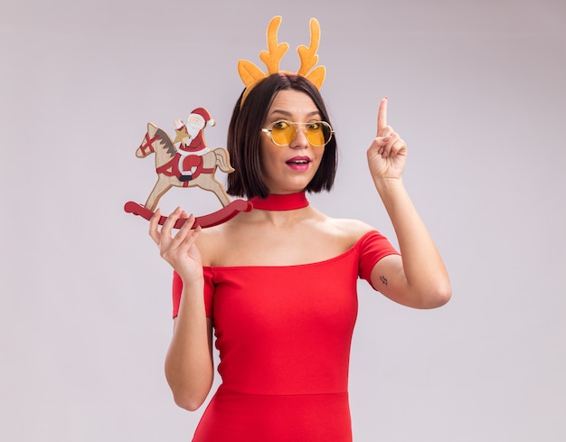 Impressed young girl wearing reindeer antlers headband and glasses holding santa on rocking horse figurine looking at camera pointing up isolated on white background