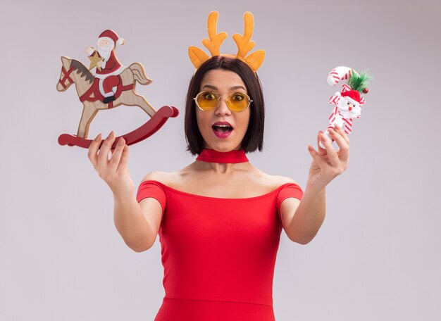 Impressed young girl wearing reindeer antlers headband and glasses stretching out santa on rocking horse figurine and candy cane ornament towards camera looking at camera isolated on white background