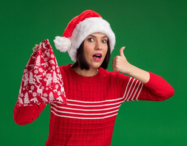 Impressed young girl wearing santa hat holding christmas gift sack looking at camera showing thumb up isolated on green background