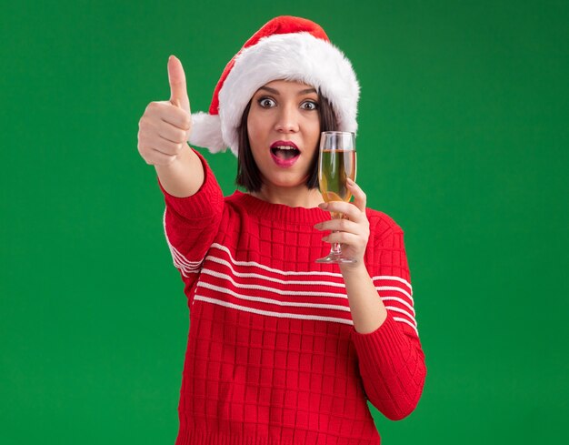 Impressed young girl wearing santa hat holding glass of champagne looking at camera showing thumb up isolated on green background