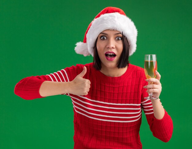 Impressed young girl wearing santa hat holding glass of champagne  showing thumb up isolated on green wall