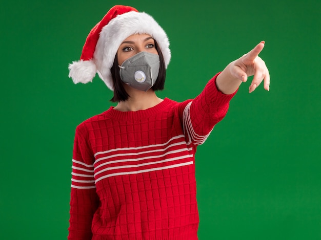 Free photo impressed young girl wearing santa hat and protective mask looking and pointing at side isolated on green wall