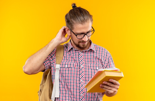 Free Photo impressed young guy student wearing backpack with glasses holding and looking at book scratching head isolated on orange wall