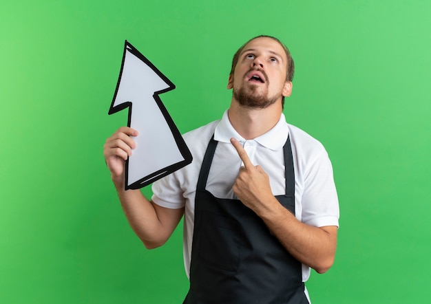 Free photo impressed young handsome barber wearing uniform holding arrow mark looking and pointing up isolated on green