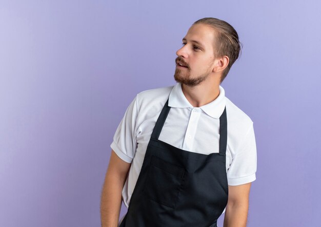 Impressed young handsome barber wearing uniform standing and looking at side isolated on purple  with copy space