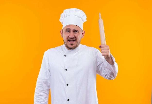 Impressed young handsome cook in chef uniform holding rolling pin isolated on orange wall