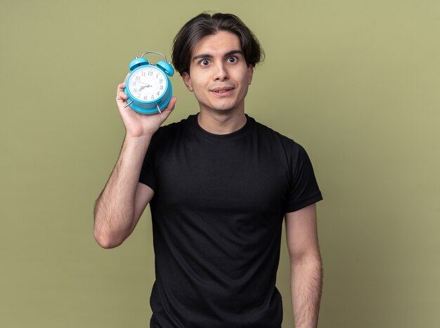 Impressed young handsome guy wearing black t-shirt holding alarm clock around face isolated on olive green wall