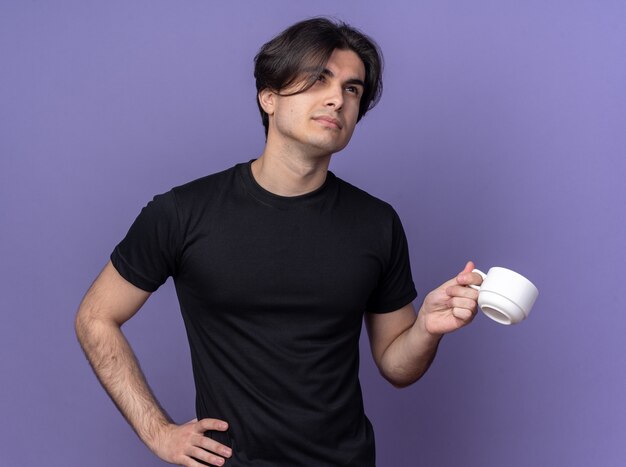 Impressed young handsome guy wearing black t-shirt holding cup of coffee putting hand on hip isolated on purple wall