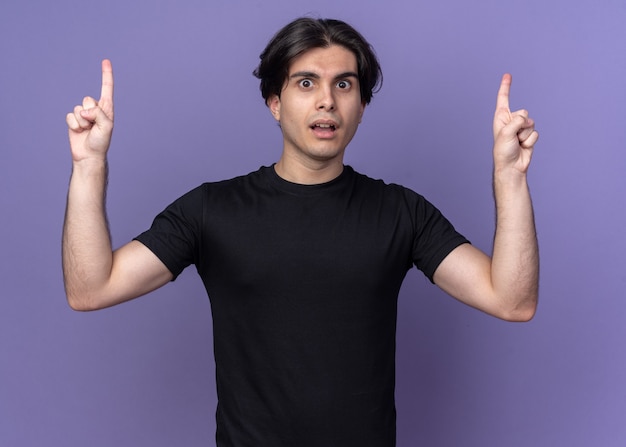 Impressed young handsome guy wearing black t-shirt points up isolated on purple wall