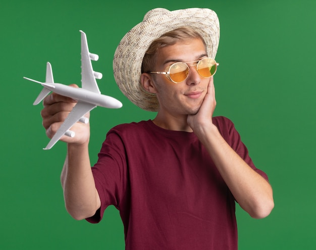 Free Photo impressed young handsome guy wearing red shirt with glasses and hat holding toy airplane putting hand on cheek isolated on green wall