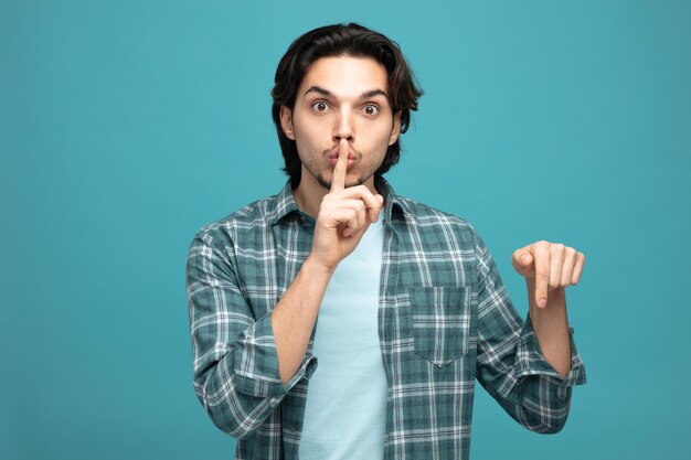 impressed young handsome man looking at camera showing silence gesture pointing down isolated on blue background